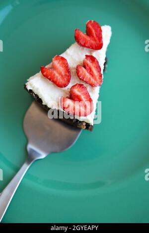 Un pezzo di cheesecake alla fragola decorato con fragole a forma di cuore giace su un piatto color menta e si erge su un tovagliolo a scacchi. Vista da abo Foto Stock