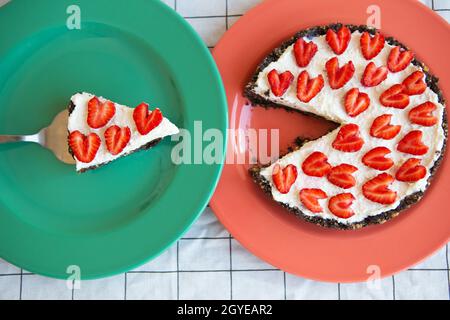 Una cheesecake alla fragola decorata con fragole a forma di cuore si trova su un piatto e si trova su un tovagliolo a scacchi, tagliare una fetta. Vista dall'alto Foto Stock