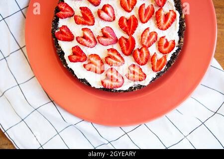 Cheesecake alla fragola decorata con fragole a forma di cuore giace su un piatto color corallo, si erge su un tovagliolo e un tavolo di legno. Immagine di una Foto Stock