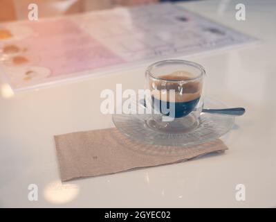 Cappuccino servito in un bicchiere trasparente a doppia parete, su un piattino con cucchiaio e tovagliolo marrone. Caffè caldo su tavola bianca. Foto Stock