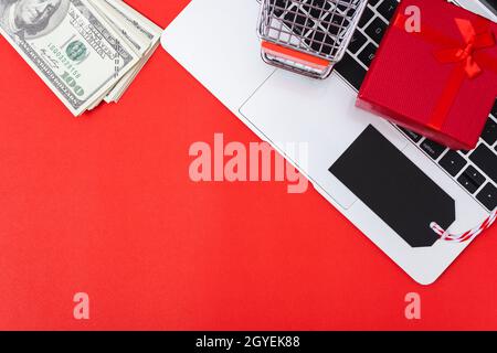 Marketing online, vista dall'alto dello spazio di lavoro con tag nero vuoto, denaro, confezione regalo e shopping carrello su computer portatile su sfondo rosso e hanno copia spa Foto Stock