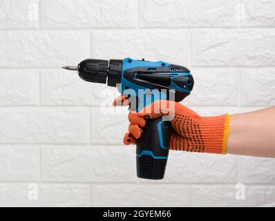 mano in un guanto di tessuto tiene un trapano portatile su una batteria contro uno sfondo di una parete di mattoni bianchi Foto Stock