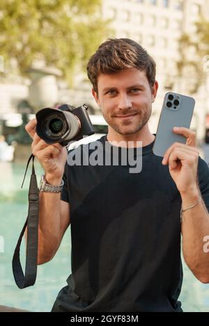 Scelta tra telefono cellulare e fotocamera DSLR per la fotografia. Uomo che tiene due fotocamere mentre si siede vicino alla fontana sulla piazza della città in giorno di sole Foto Stock