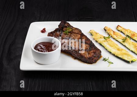 Bistecca di manzo nero angus alla griglia a New York con zucchine su piatto bianco su sfondo nero di legno con Foto Stock