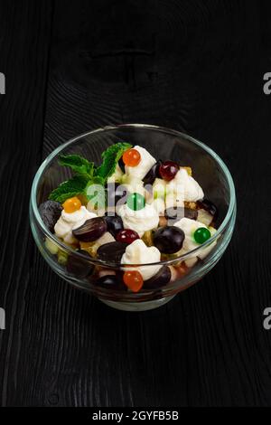 Insalata di uva mele pere arance kiwi con mascarpone chease e crema. Insalata estiva di frutta fresca sana in una ciotola di vetro su un backgroun di legno nero Foto Stock