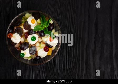 Insalata di uva mele pere arance kiwi con mascarpone chease e crema. Insalata estiva di frutta fresca sana in una ciotola di vetro su un backgroun di legno nero Foto Stock