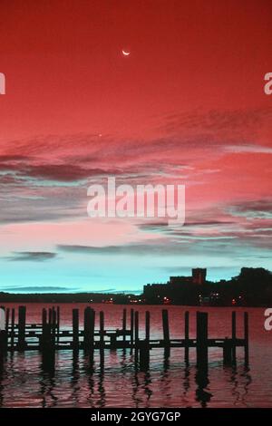La silhouette infrarossa di banchine vuote in un fiume viola e teale con un cielo rosso e viola con una luna crescente e nuvole blu cielo all'alba Foto Stock