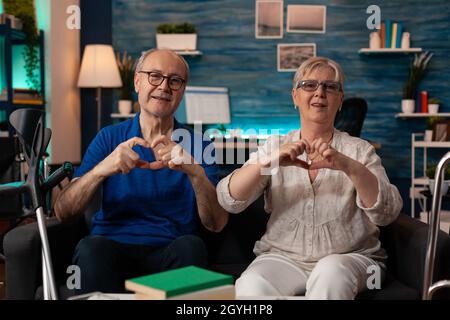 Coppia anziana che crea la forma del cuore con le mani mentre guarda la fotocamera in salotto. Persone sposate in amore che fanno insieme simbolo di affetto e che si siedono sul divano a casa. Rapporti senior Foto Stock