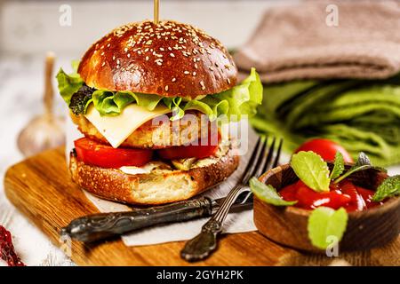 Hamburger di pollo appetitoso fatto in casa con formaggio, insalata fresca e verdure su un tagliere. Foglie di lattuga fresca, pomodori e cipolle. Tradizionale Foto Stock