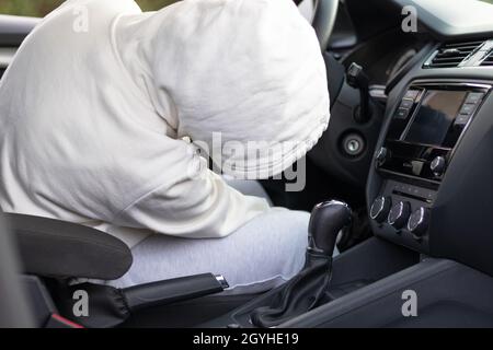 Un uomo in un maglione bianco con un cappuccio siede al volante di una macchina e cerca di avviarla per rubarla. Messa a fuoco selettiva. Primo piano Foto Stock