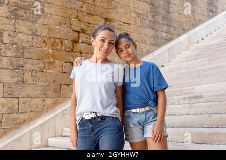 Madre di mezza età e sua figlia bambino in piedi su scale di pietra all'aperto, abbracciando e guardando la macchina fotografica. Famiglia amorevole che indossa magliette blu e jea Foto Stock