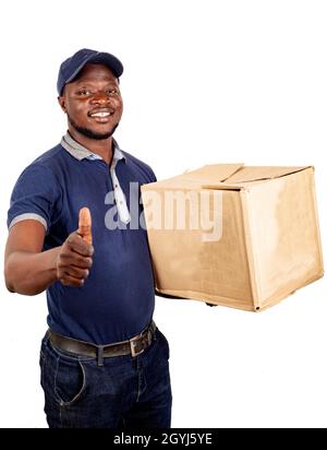 Il giovane addetto alle consegne, in piedi in uniforme su sfondo bianco, con una grande scatola di cartone, guarda la fotocamera facendo gesti con il pollice sorridendo. Foto Stock