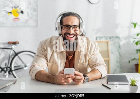 entusiasta freelancer ispanico in cuffie ridendo alla fotocamera mentre ascolti il podcast a casa Foto Stock