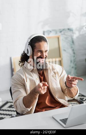 entusiasta freelance latino nelle cuffie che puntano al notebook durante le videochiamate a casa Foto Stock