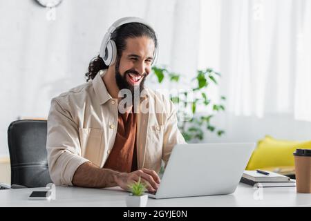 ridendo il freelance ispanico in cuffie digitando sul portatile mentre si lavora a casa Foto Stock
