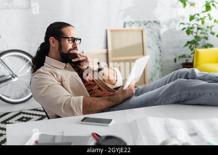 design latino attento che guarda il tablet digitale mentre sei seduto con le gambe sulla scrivania vicino a uno smartphone sfocato Foto Stock
