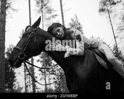 Un giovane e bellissimo pilota di brunette in un elegante abito retrò seduto a cavalcare un cavallo nero appoggiato sul collo. Tinta bianca e nera Foto Stock