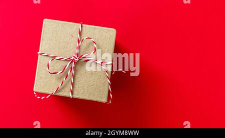 Confezione regalo con arco legato a righe rosso Natale bianco, regalo vacanza kraft marrone su sfondo rosso, vista dall'alto. Confezionatrice fai da te ecologica fatta in casa Foto Stock