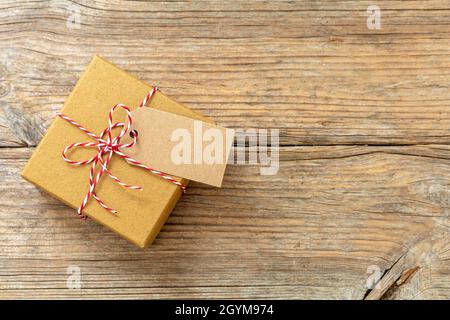 Scatola regalo di Natale con scheda di nota vuota, regalo di festa di carta kraft marrone con arco legato a strisce rosso bianco su sfondo tavolo di legno, vista dall'alto Foto Stock