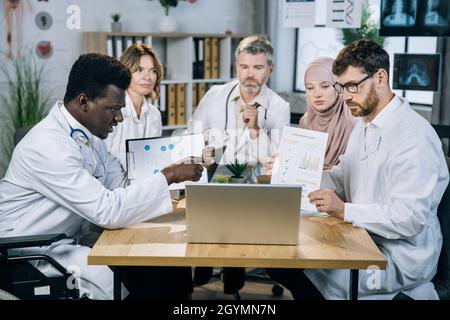 Cinque specialisti medici multietnici che mostrano vari grafici e grafici durante le videochiamate su un moderno notebook. Gruppo di medici competenti che hanno una conferenza online in ufficio. Foto Stock