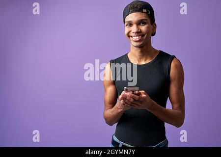 Uomo sorridente transgender con cellulare in mano su sfondo viola. Uomo allegro in abito casual Foto Stock