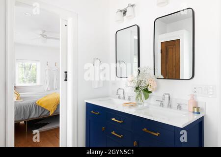 Un bel bagno rinnovato con un vanity blu, piano di appoggio in marmo bianco e specchi neri che guardano verso una camera da letto. Foto Stock