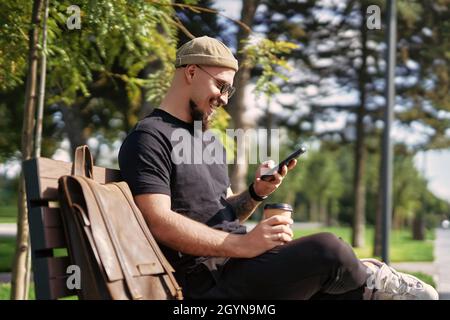 Vista laterale di uomo sorridente indossare abiti casual sedersi sulla panca mentre surf smartphone all'aperto in strada Foto Stock