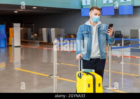 Uomo in maschera medica viso utilizzando smartphone dietro il banco check-in all'interno dell'aeroporto durante il blocco covid. Il viaggiatore informale urbano ha un telefono cellulare Foto Stock