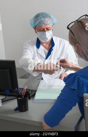 Un medico in un cappotto bianco, maschera e cappuccio in un ufficio medico seduto a un tavolo dà una penna a un paziente per firmare un consenso informato Foto Stock