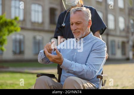 Uomo anziano in sedia a rotelle pulendo le mani con disinfettante antibatterico come misura igienica per prevenire l'infezione Covid19 mentre cammina assistito da Foto Stock