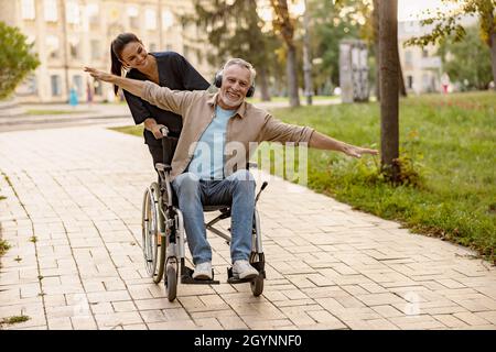 Gioioso uomo disabili maturo in sedia a rotelle indossando le cuffie divertirsi durante una passeggiata in città assistita da una bella giovane infermiera Foto Stock