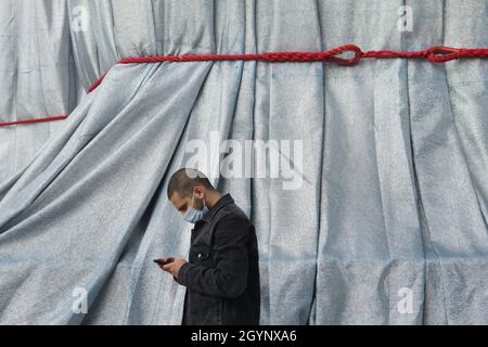 Il giovane che indossa una maschera usa uno smartphone davanti all'Arco di Trionfo avvolto in tessuto argento-blu con corde rosse in Place Charles de Gaulle a Parigi, Francia. L'Arco di Trionfo è stato avvolto per due settimane convertendosi in un'opera d'arte come è stato progettato da Christo e Jeanne-Claude nel settembre 2021. Foto Stock