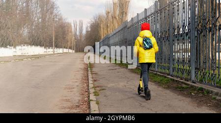 Il bambino sta guidando uno scooter. Una ragazza in una giacca gialla guida fuori in distanza su uno scooter su una strada vuota lungo una recinzione di ferro. Spazio di copia Foto Stock