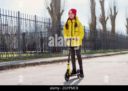 Una ragazza in una giacca gialla che guida uno scooter su una strada vuota. Concetto di trasporto ecologico e per bambini. Foto Stock