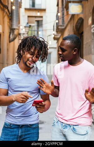 attraente giovane uomo latino-americano a piedi strade con un amico africano. Stanno parlando mentre controllano uno smartphone rosso Foto Stock