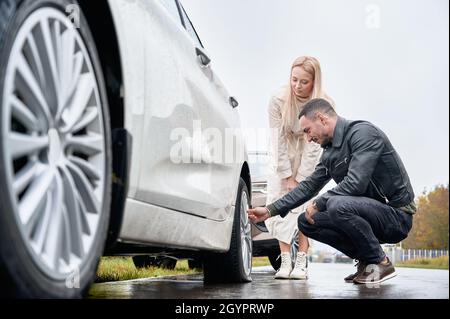 Bella giovane donna in piedi accanto all'automobile e sorridente mentre l'uomo controlla la ruota. Bel meccanico auto che aiuta il conducente femminile a cambiare pneumatico auto sulla strada. Foto Stock