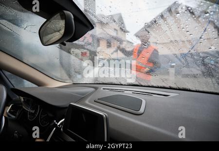 Vista ravvicinata dall'interno dell'auto attraverso il parabrezza bagnato. Su sfondo sfocato auto lavaggio specialista in uniforme luminosa con pistola ad alta pressione in mani, edifici di appartamenti. Foto Stock