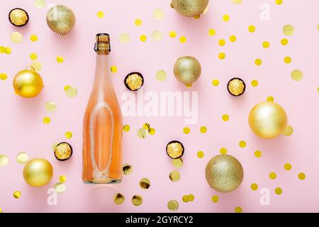 Bottiglia di champagne rosa con confetti, caramelle dolci e palle di Natale decorazioni natalizie dorate su sfondo rosa. Disposizione piatta Foto Stock