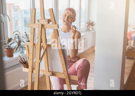 Donna positiva con capelli colorati morsi matita seduta a cavalletto Foto Stock