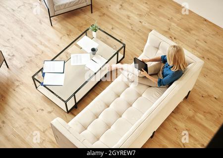 Vista ad alto angolo di una donna matura in abbigliamento casual utilizzando un tablet pc mentre si lavora da casa, si fa lavoro di carta, seduto su un divano in un appartamento moderno Foto Stock