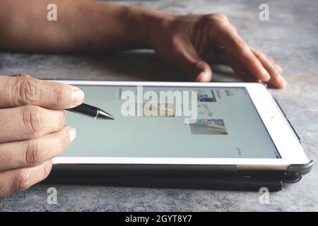 Primo piano della mano destra di un uomo d'affari che tiene una penna nera su un tablet sfocato con luce arancione al mattino per utilizzare i social media o fare una pausa Foto Stock
