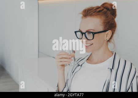 Sorridente ragazza rossa tiene un bicchiere di acqua beve acqua per rinfrescarsi il corpo Foto Stock