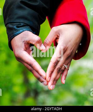 le mani nere della coppia che formano il cuore in giacche rosse e nere nella foresta Foto Stock
