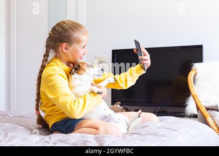 Una ragazza teen con un telefono cellulare in mano è blogging circa gli animali domestici, seduta sul letto con un cane nelle sue braccia. Un bambino è fotografato o prende un Foto Stock