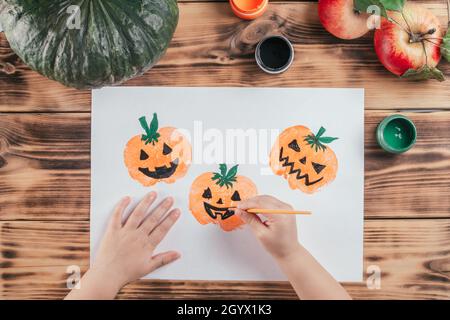 Passo-per-passo Halloween bambini tutorial stampe di mela zucca. Punto 10: La mano del bambino disegna gli occhi e la bocca delle zucche con vernice nera del gouache. Vista superiore Foto Stock