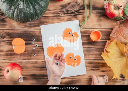 Passo-per-passo Halloween bambini tutorial stampe di mela zucca. Passo 11: La mano del bambino tiene gli occhi per incollare alla stampa della zucca. Vista dall'alto Foto Stock