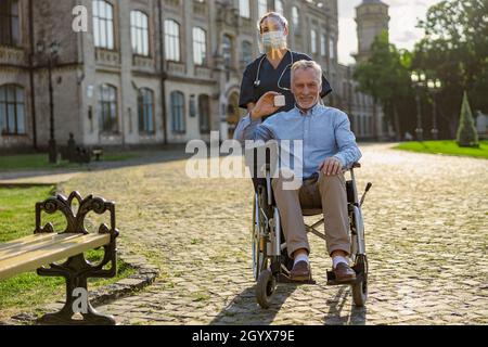 Scatto a tutta lunghezza di uomo anziano felice che recupera il paziente che tiene il vaso di crema, sorridendo alla macchina fotografica su una passeggiata all'aperto vicino alla clinica di riabilitazione Foto Stock