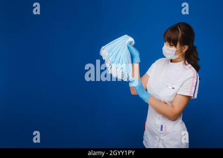 Una giovane ragazza medico in una maschera medica tiene un sacco di maschere nelle sue mani. Un'infermiera con maschere bianche nelle mani su sfondo blu. Foto Stock