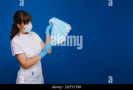 una ragazza medico in una maschera medica su sfondo blu tiene un sacco di maschere protettive nelle sue mani. Foto Stock