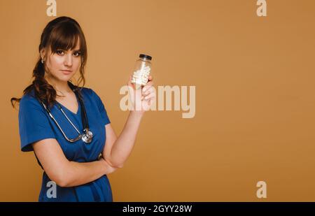 Un medico in un camice da laboratorio blu tiene un vaso di capsule nelle sue mani su uno sfondo marrone. Foto Stock